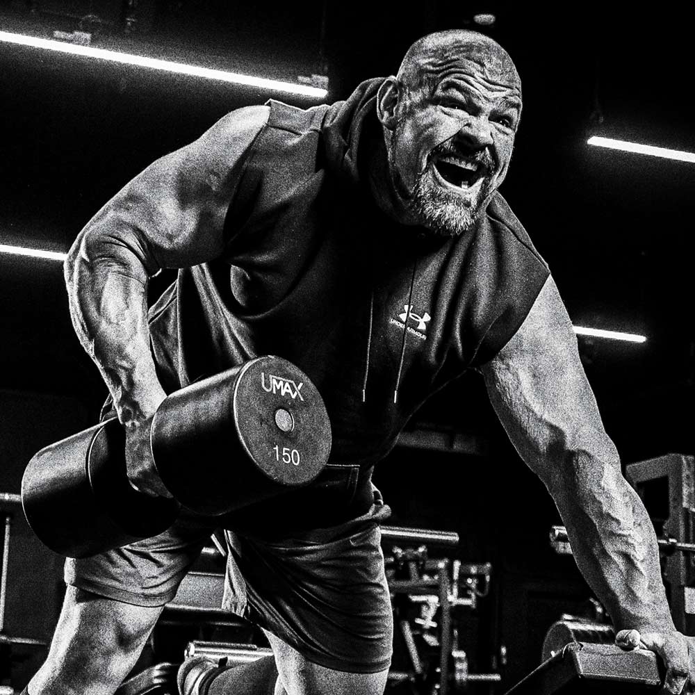 Brian Shaw in a gym lifting a 150 lb dumbbell, wearing a sleeveless Under Armour hoodie and shorts. The photo is in high-contrast black and white, with bright gym lights in the background.