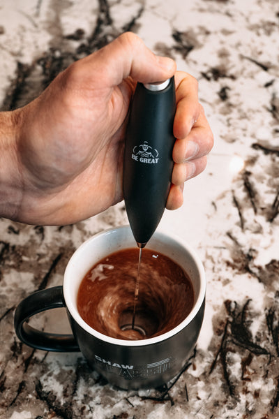 A hand holding a Shaw Strength Be Great frother over a mug filled with a chocolate beverage, on a kitchen counter top.