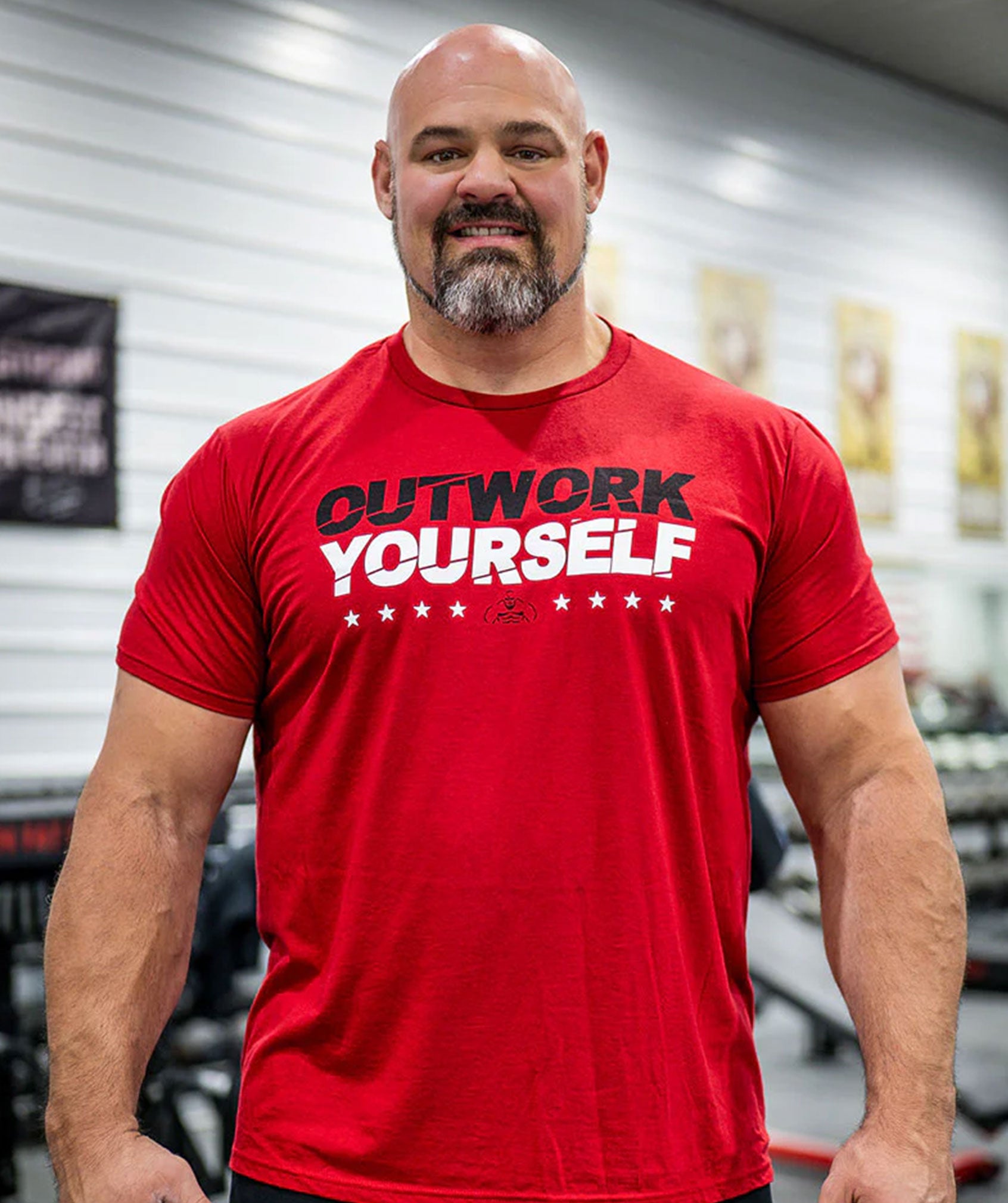 Brian Shaw wearing a red t-shirt with 'OUTWORK YOURSELF' text in a gym setting