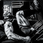A black and white photograph of Brian Shaw lying on a gym leg press machine, wearing a sleeveless shirt and shorts, with his legs extended toward a pair of shoes in the upper right.
