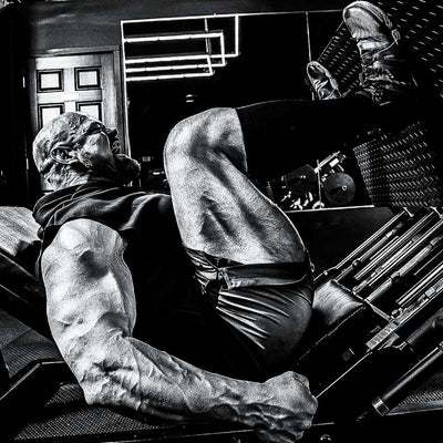 A black and white photograph of Brian Shaw lying on a gym leg press machine, wearing a sleeveless shirt and shorts, with his legs extended toward a pair of shoes in the upper right.