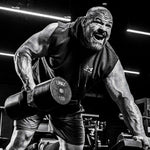 Brian Shaw in a gym lifting a 150 lb dumbbell, wearing a sleeveless Under Armour hoodie and shorts. The photo is in high-contrast black and white, with bright gym lights in the background.