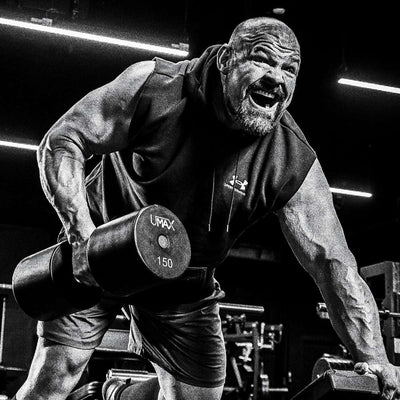 Brian Shaw in a gym lifting a 150 lb dumbbell, wearing a sleeveless Under Armour hoodie and shorts. The photo is in high-contrast black and white, with bright gym lights in the background.
