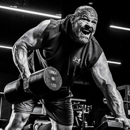 Brian Shaw in a gym lifting a 150 lb dumbbell, wearing a sleeveless Under Armour hoodie and shorts. The photo is in high-contrast black and white, with bright gym lights in the background.