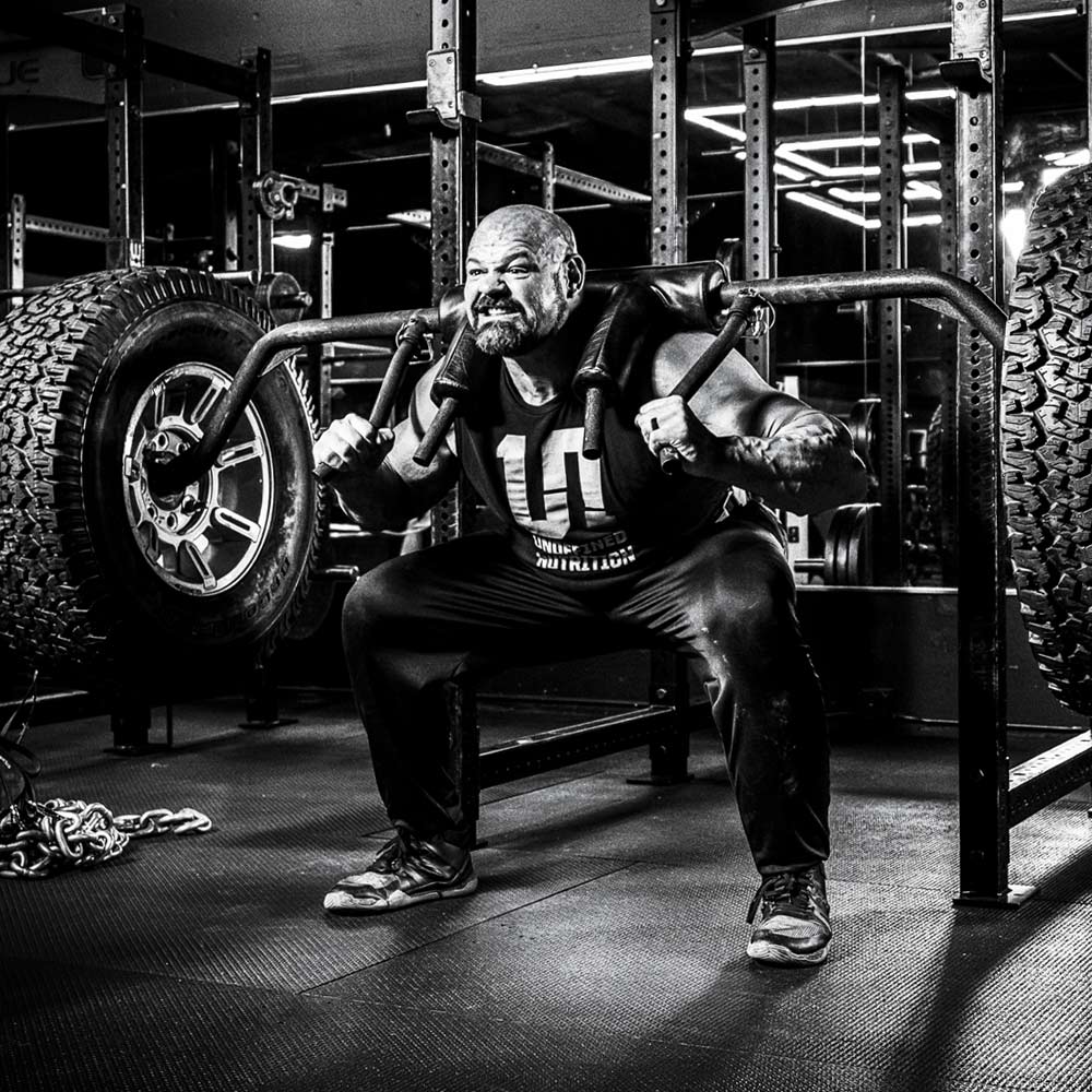 A black and white photograph of Brian Shaw in a gym, performing a squat on a bench with a barbell across his upper chest. He is wearing a dark t-shirt, dark sweatpants, and athletic shoes.
