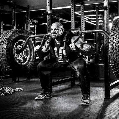 A black and white photograph of Brian Shaw in a gym, performing a squat on a bench with a barbell across his upper chest. He is wearing a dark t-shirt, dark sweatpants, and athletic shoes.