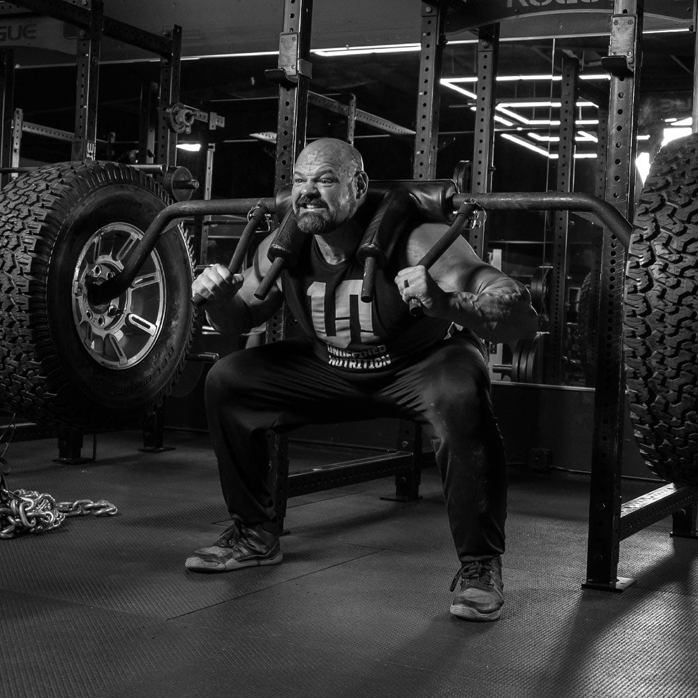 Brian Shaw in a gym wearing a sleeveless athletic shirt and squatting on under a barbell rack with large tires.