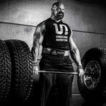 Brian Shaw in a black sleeveless tank top with 'UNDEFINED NUTRITION' printed on the front, lifting a barbell on a gym floor between large tires. He is wearing a belt with the Evolution Athletics lifting belt.