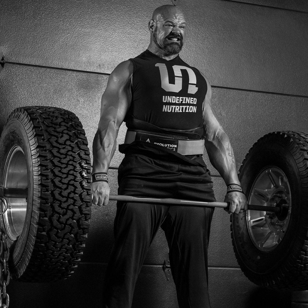 Brian Shaw in a black sleeveless tank top with 'UNDEFINED NUTRITION', lifting a barbell loaded with large off-road tires. The photo is in black and white, set against a textured wall.