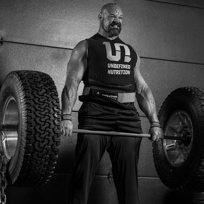 Brian Shaw in a black sleeveless tank top with 'UNDEFINED NUTRITION', lifting a barbell loaded with large off-road tires. The photo is in black and white, set against a textured wall.