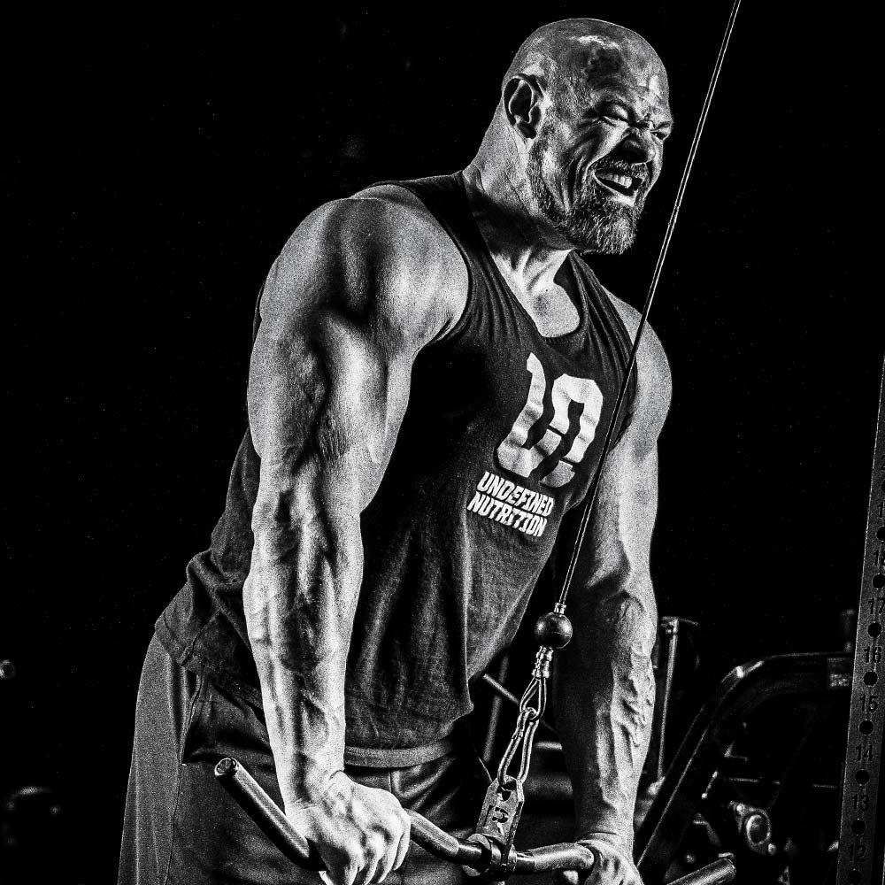 Brian Shaw in a black tank top with 'UNDEFINED NUTRITION' and a large number 10 printed on the front, performing a cable pull-through exercise in a gym setting. The photo is in high-contrast black and white.