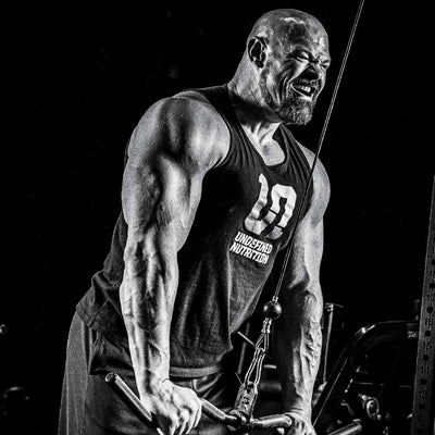 Brian Shaw in a black tank top with 'UNDEFINED NUTRITION' and a large number 10 printed on the front, performing a cable pull-through exercise in a gym setting. The photo is in high-contrast black and white.
