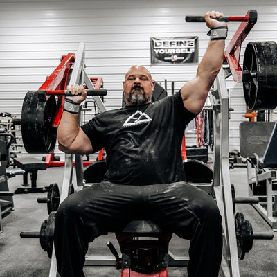 Brian Shaw performing a bench press in a gym setting with weights and gym equipment around.