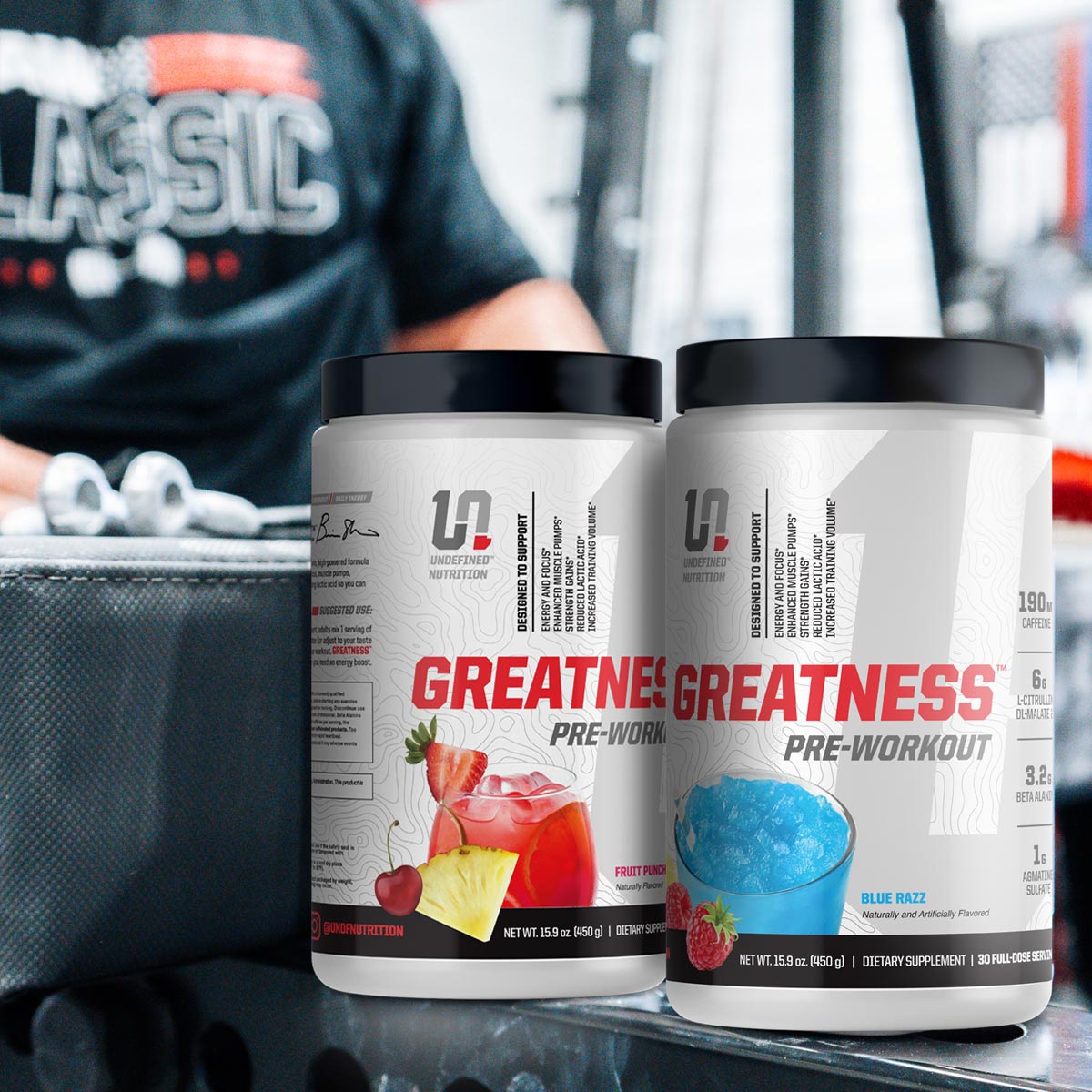 Two containers of 'Greatness' pre-workout supplement with a person in the background.