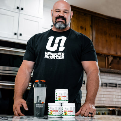 Brian Shaw wearing a black 'Undefined Nutrition' t-shirt with nutritional supplements on a kitchen counter.