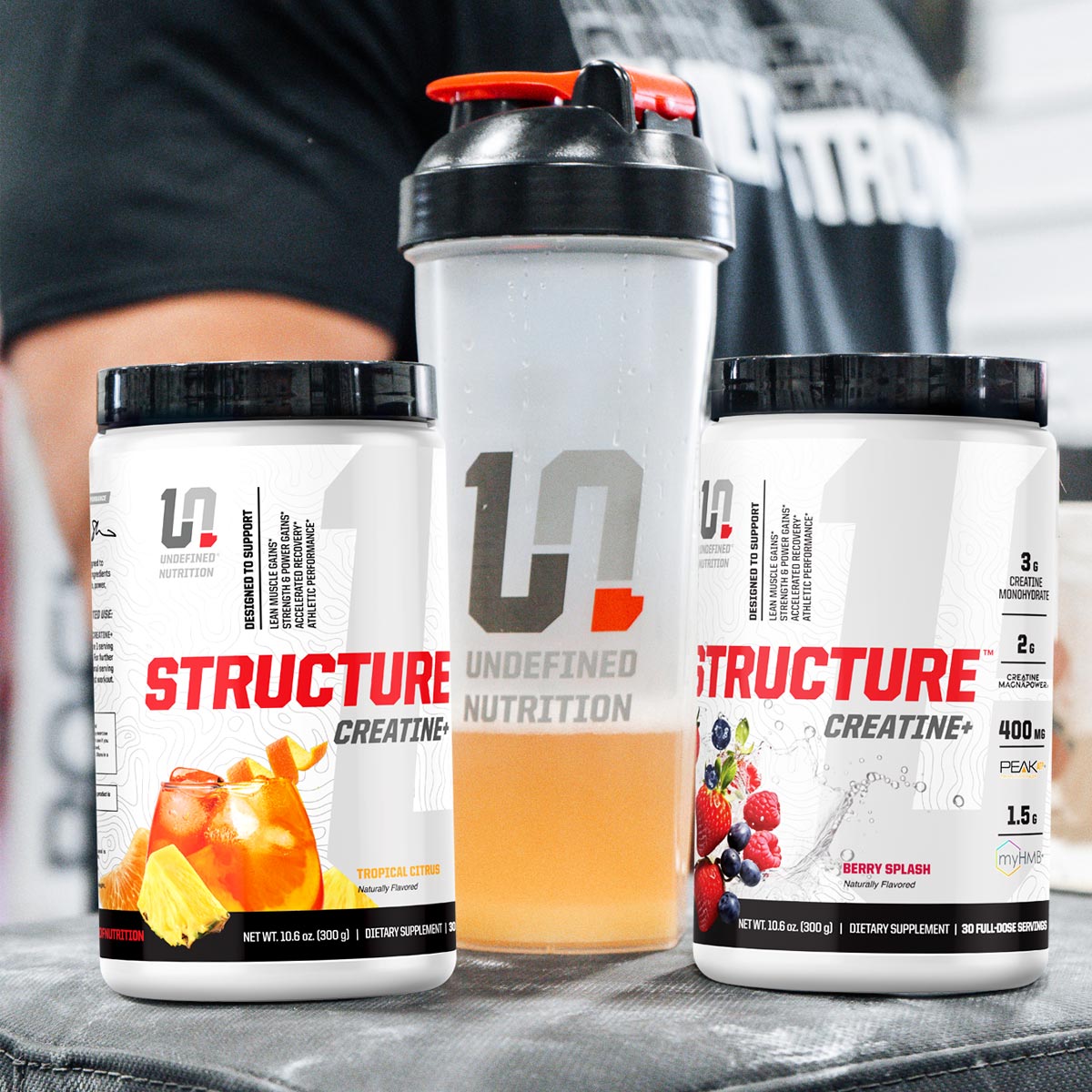 Two containers of Structure Creatine+ and a shaker bottle on a surface with a person in the background.